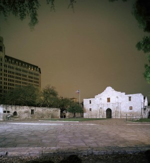 a view of a stone paved plaza with a two story white stone building with a central arched doorway. In the background is a multi floored flat iron style skyscraper and a murky night sky. 