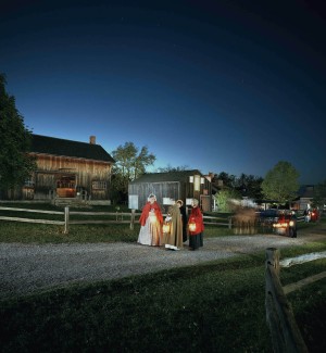 a nightime outdoor setting in an historic rural village with three women in long full dresses and bonnets holding lanterns. Worn wooden sided barns and split rail fences in the background. 