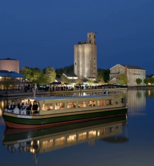 a view of a long wooden river boat with a roof carrying passengers standing on the ooen deck and inside throuh the windows. The boat is reflected in th canal water and a series of old mill building are on the opposite shore with a deeep blue nighttime sky. 