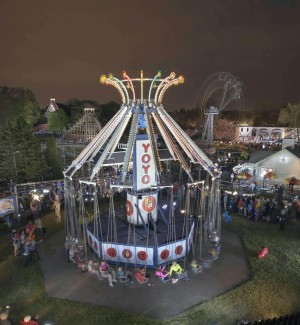 panoramic view of an amusement park with a large cicular swing ride with 'YOYO' in lights at the top of the roof. A wooden roller coaster and ferris wheel are in the background against a chocolate brown sky.