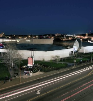 a color photograph of a nightime view of a city streetscape with a road in the foreground and a low white boxy building with a sculptural portal entry.