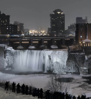 a color photograph of a winter and nighttime view of a river with frozen waterfall with a city scape with highrise buildings in the background.