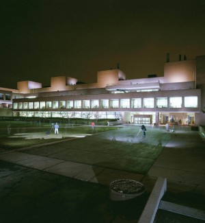 a panoramic nightime color photograph of along red brick building with a ribbon of windows lit up internally.