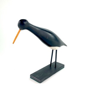 a carved wood bird form painted black with a white belly and yellow beak on a black block stand.
