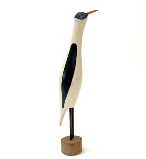 a carved wood bird form standing tall and sleek painted with a white body, black wings and yellow beak standing on a naturl round of wood with the bark.