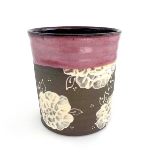 A cylindrical brown tone ceramic vessel with a rose colored rim and a decorative illustration of roses in white. 