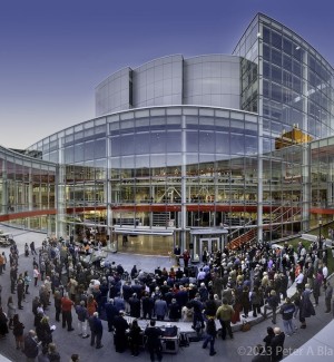 a panoramic view of a glass and steel clad modern building with an expansive walkway filled with a crowd of a hundred people gathered to watch a ribbon cutting. 