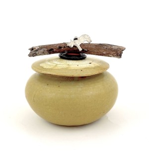 an ochre colored round ceramic jar with a lid embellished with piece of driftwood and metal charm in the shape of a cow.