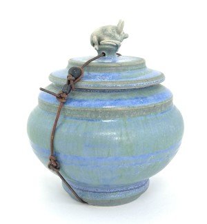 A blue ceramic lidded jar with a small sculpture of a bat on top, tied with a waxed leather cord. 