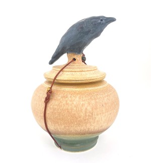 A peach hued ceramic lidded blue jar with sculpted black crow on top and a waxed leather cord tied around it.