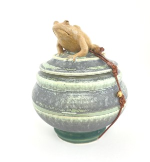 a round blue-green ceramic pot with a lid add a sculpted brown toad perched on top. 