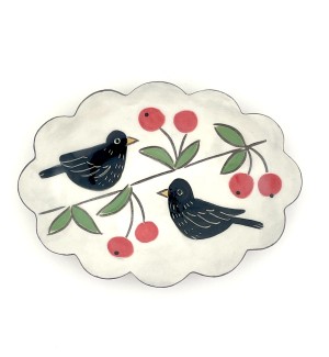 a ceramic trinket tray with a fluted edge with a white background and an illustration of a black bird perched on a branch with red cherries.