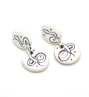 a pair of white ceramic dangle earrings with a round and oval disc and illustrations of a serpent surrounded by small red hearts.