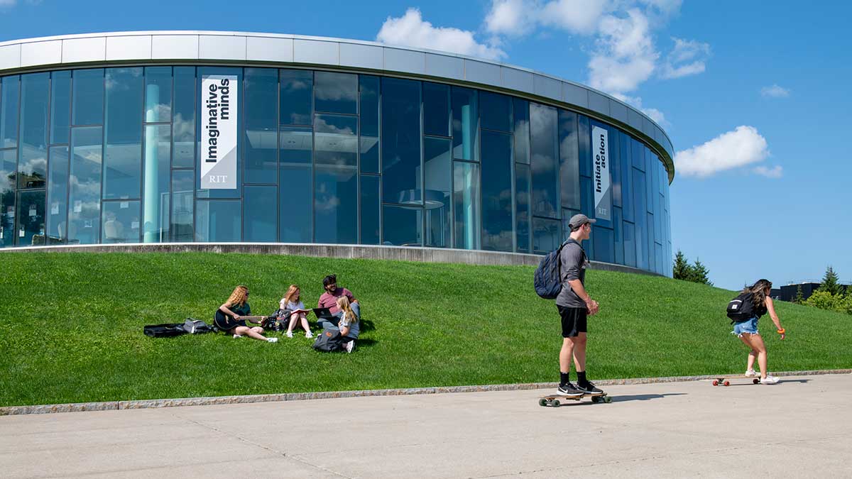 Students sit on a grassy hill outside a modern glass building while others ride skateboards on the sidewalk.