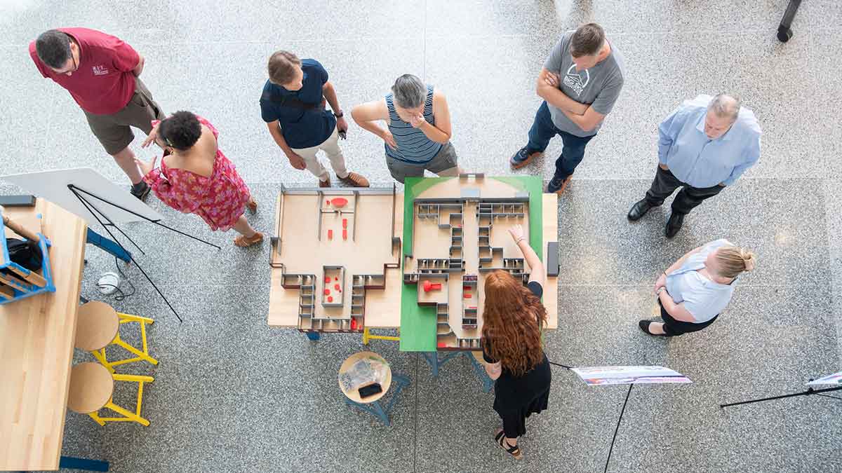 A group of people observe and discuss architectural models on tables in a design studio.