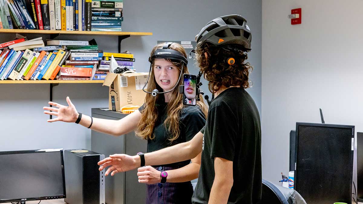 Two students wearing head-mounted devices interact in a lab surrounded by books and computer equipment.