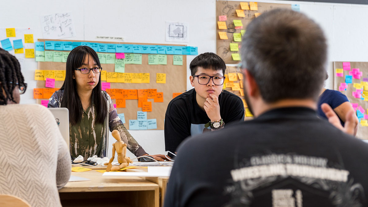 Students gather around a table covered in sticky notes and brainstorming tools during a collaborative workshop.