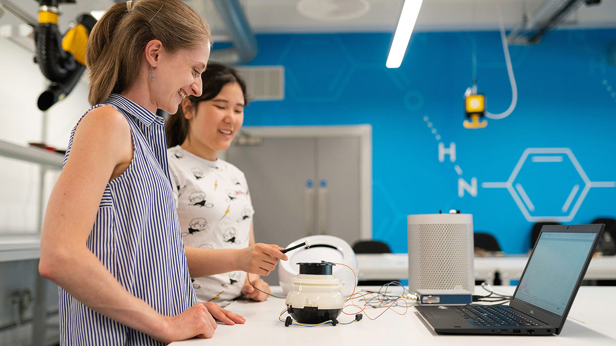 Two women smile and interact with a small electronic prototype device in a brightly lit lab with a blue chemistry-themed wall.