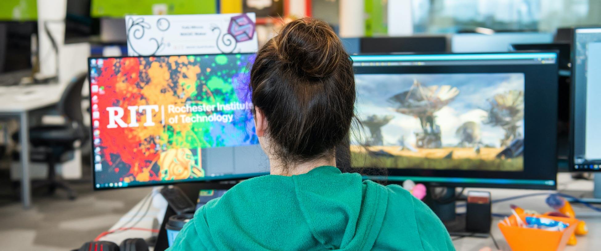 Student wearing green hoodie sites in front of two monitors.