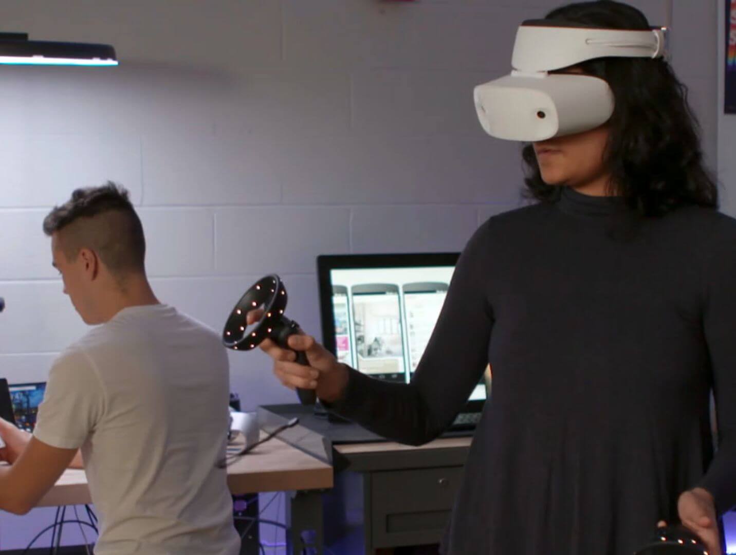A person using a virtual reality headset and controllers while another person sits in the background.