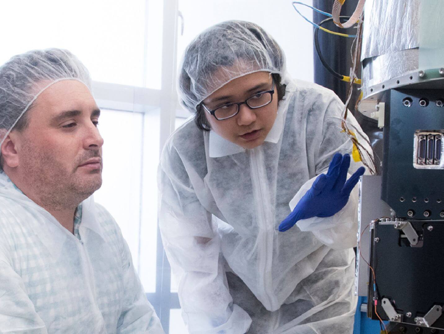 A professor and student in clean suits looking at a piece of scientific equipment.