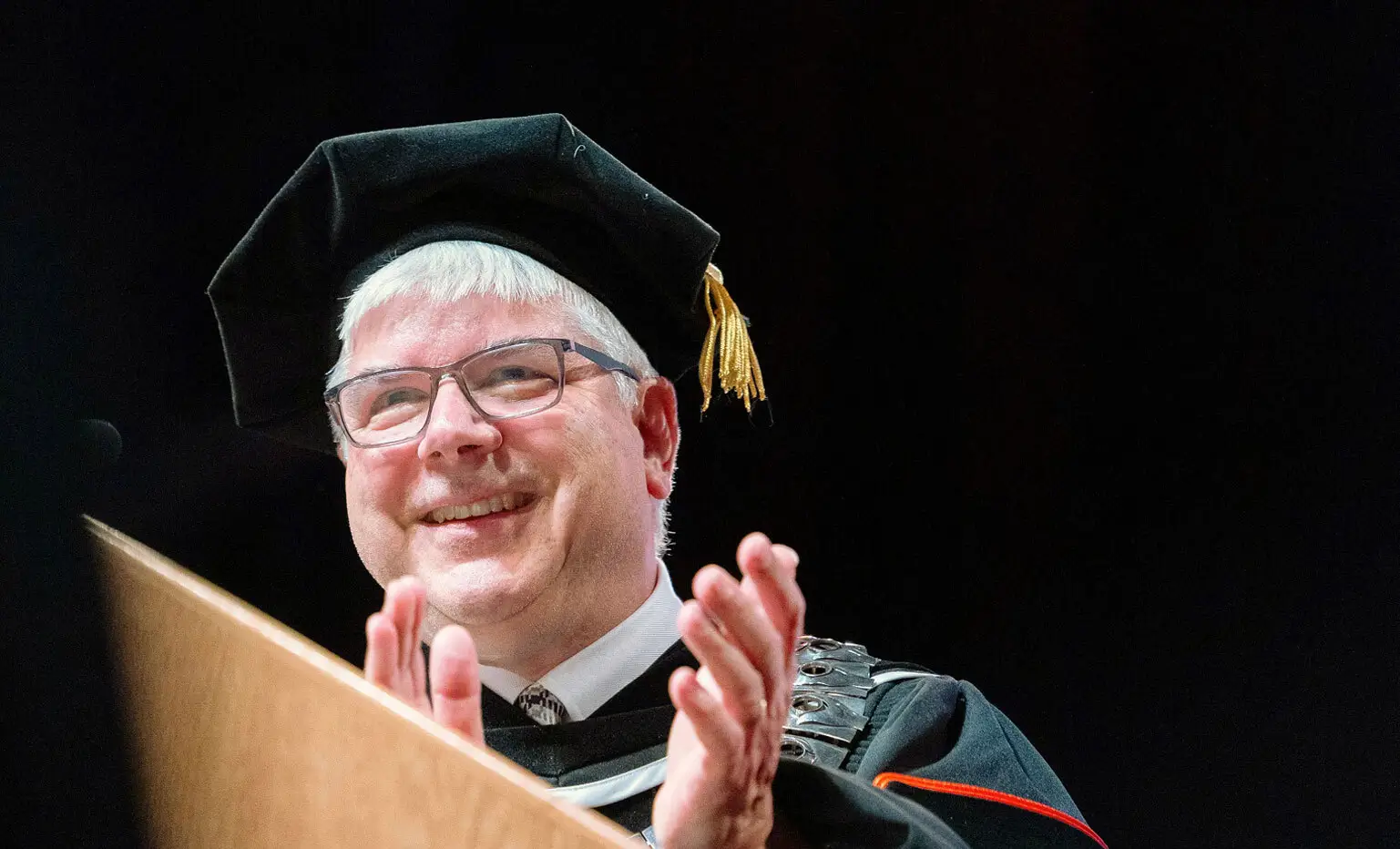 President Sanders, dressed in academic regalia, claps behind a podium.