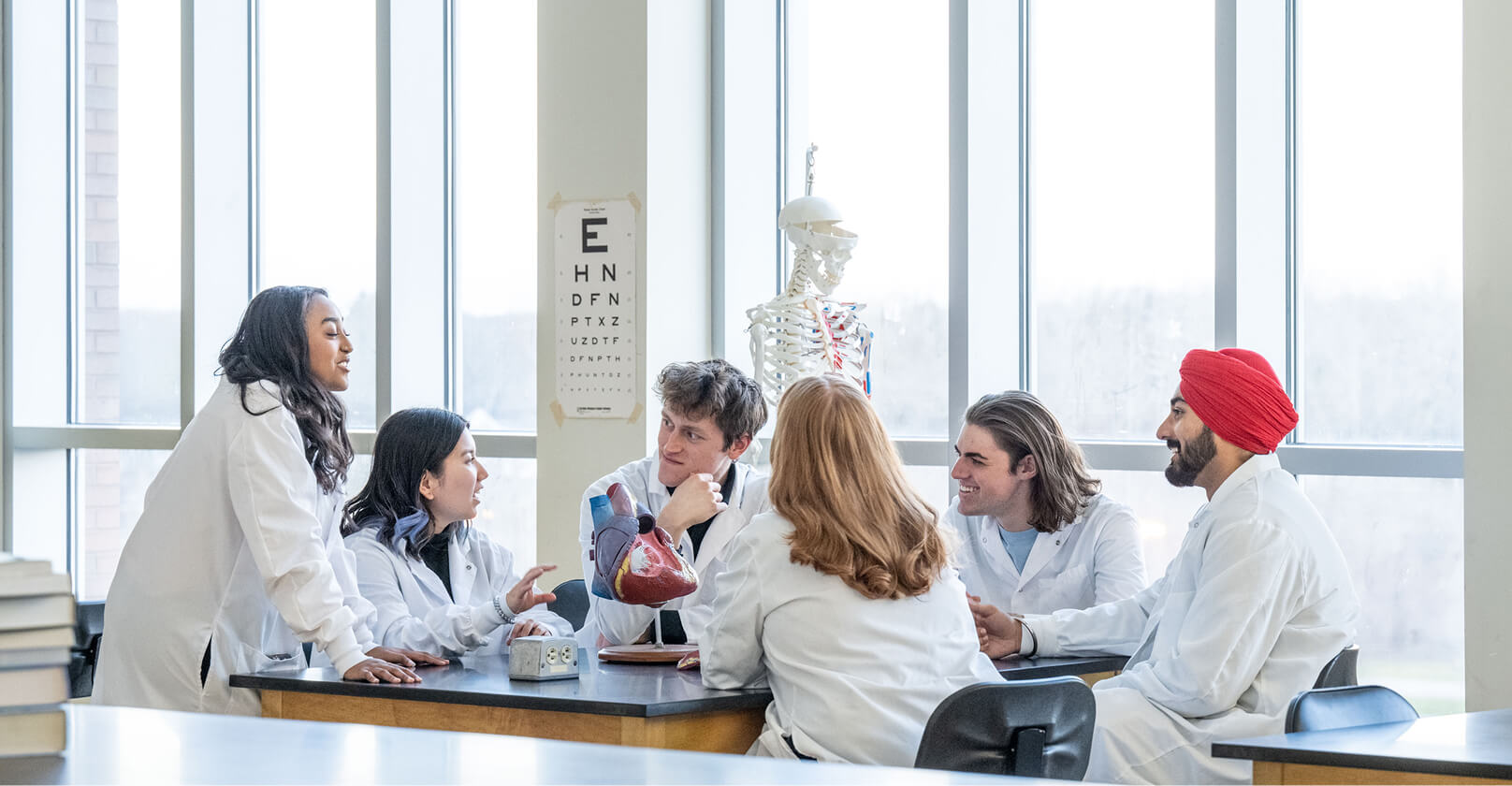 Six students wearing lab coats gathered around a table with a large model of a heart in the center.