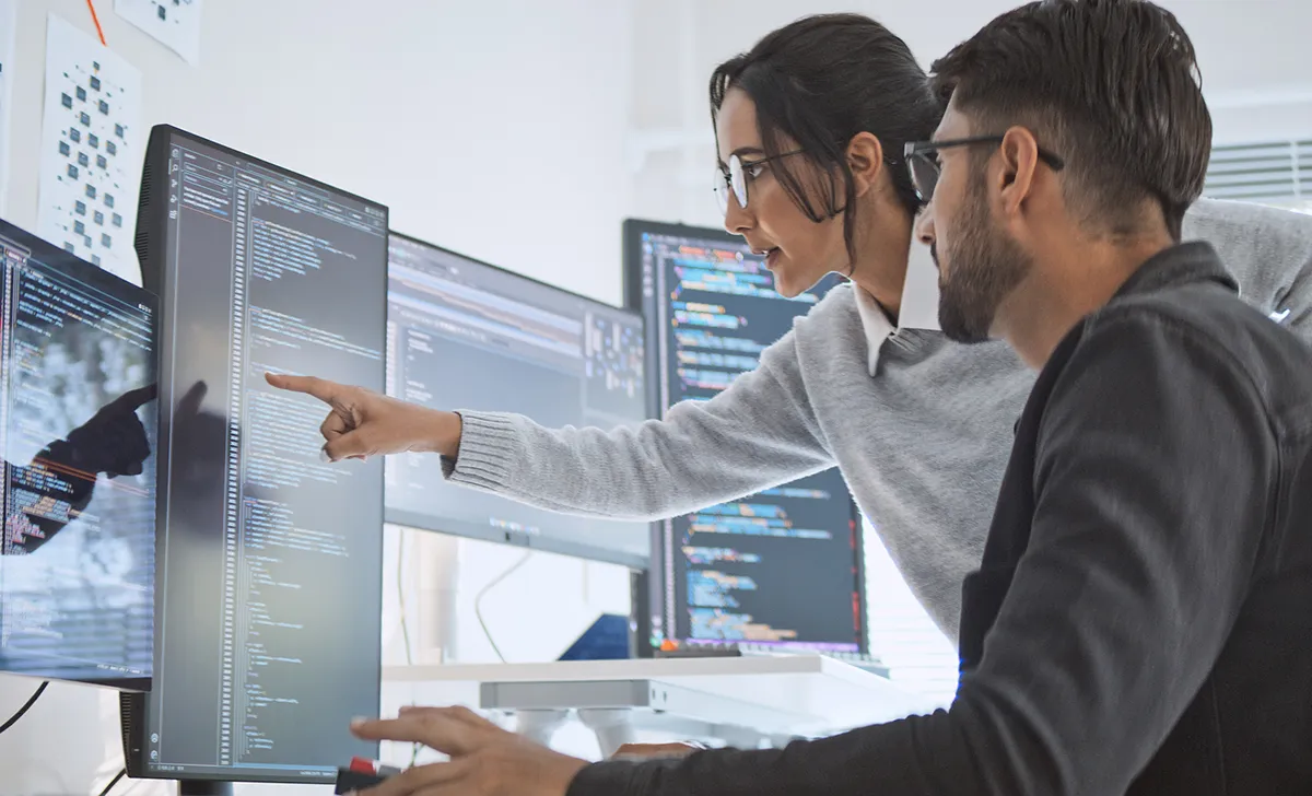 Two coworkers review code on multiple monitors, with one person pointing to a section on the screen while the other looks on.