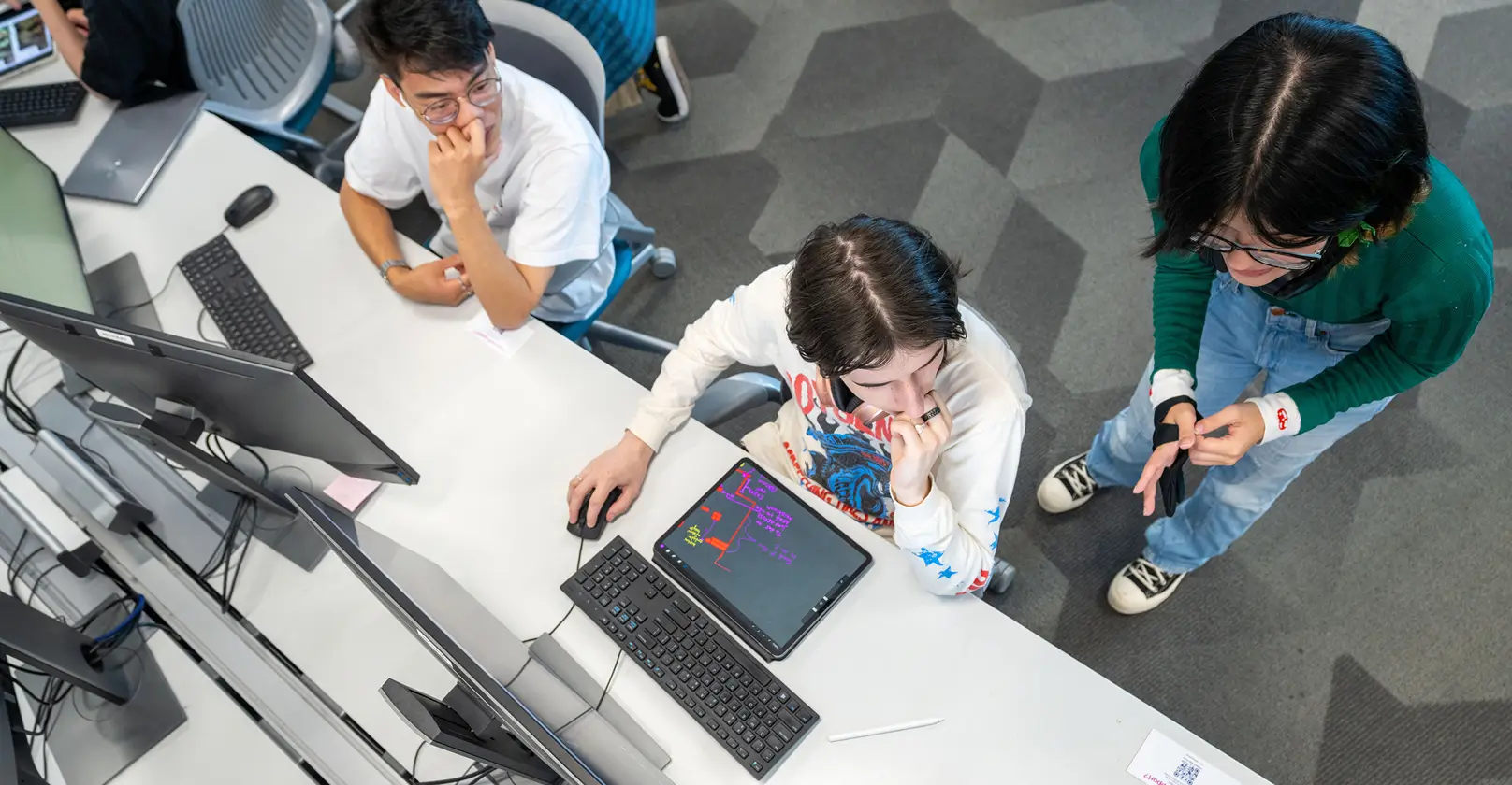 Aerial view of two people sitting in front of computers, with one person standing next to them.