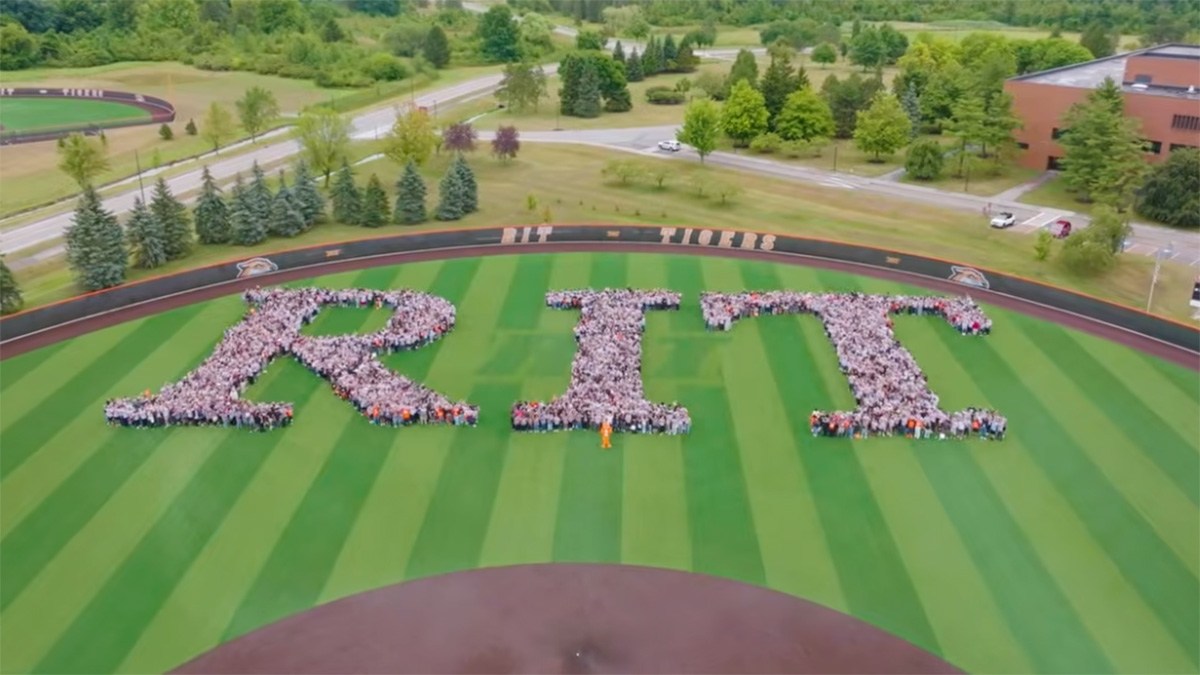 People spelling out the letters R I T on a sports field with green stripes of grass.