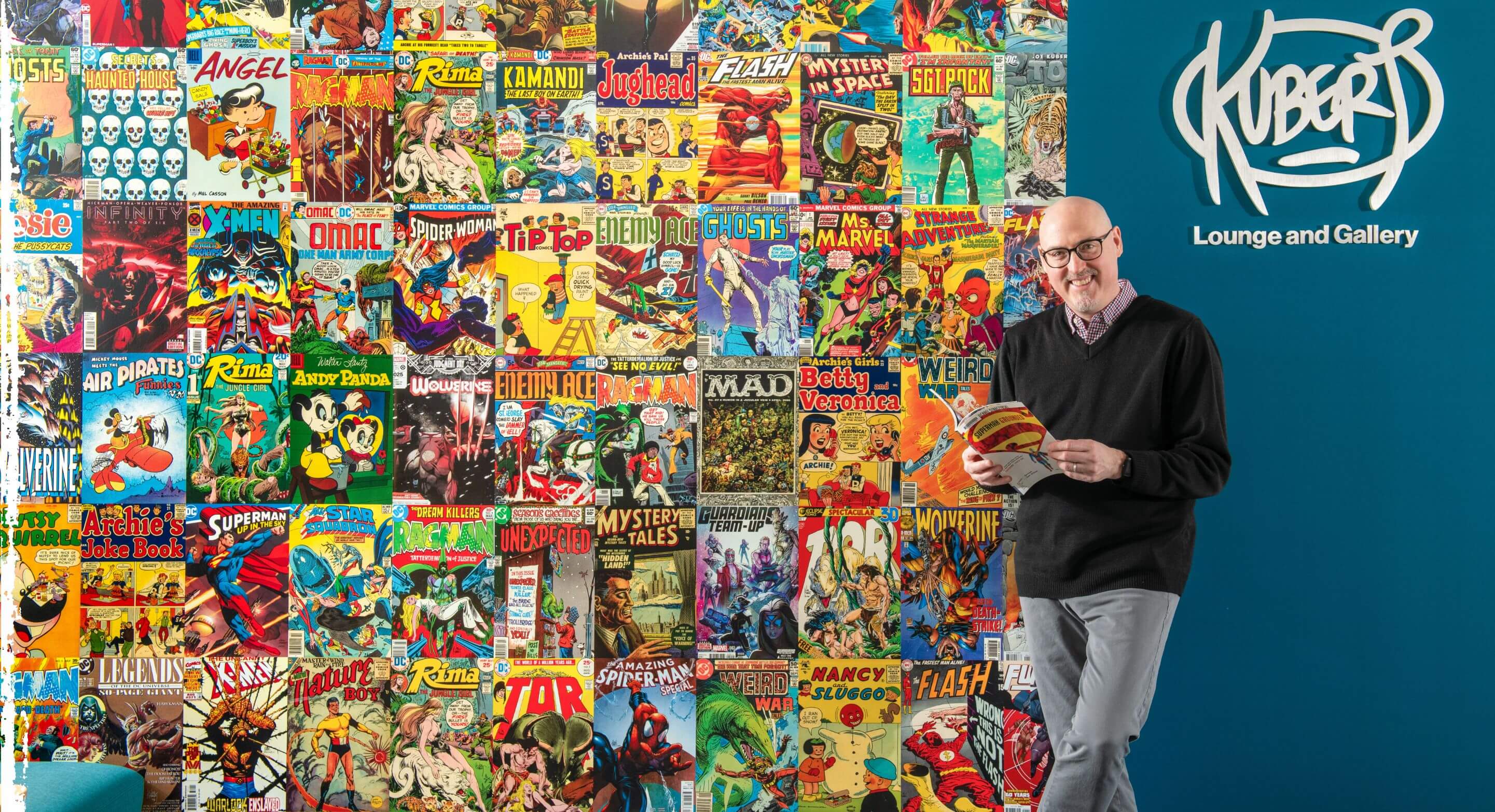 Man holding a comic book leaning against a wall with a collage of varying comic book covers and a sign that reads, Kubert Lounge and Gallery.