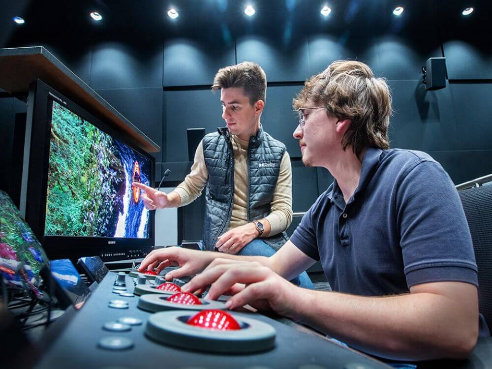 Two people working at a workstation, one points to the monitor while the other makes changes.