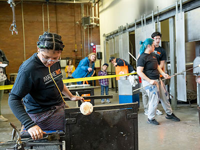 Student blows glass in foreground with students at the kiln in the 
background.