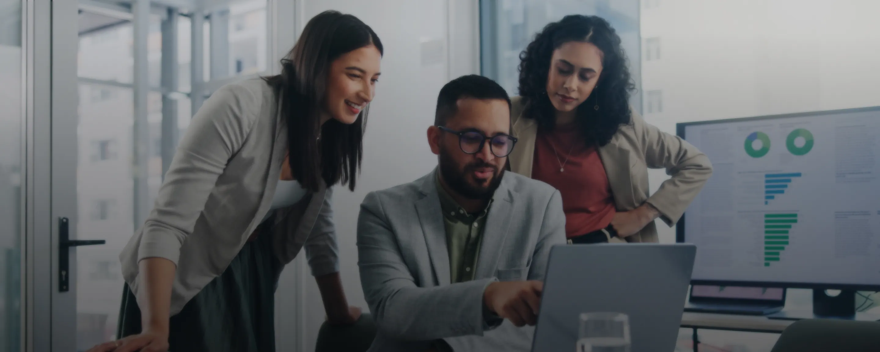 Three coworkers review information on a laptop in an office, with two people standing behind one seated person. A monitor in the background displays charts and graphs.
