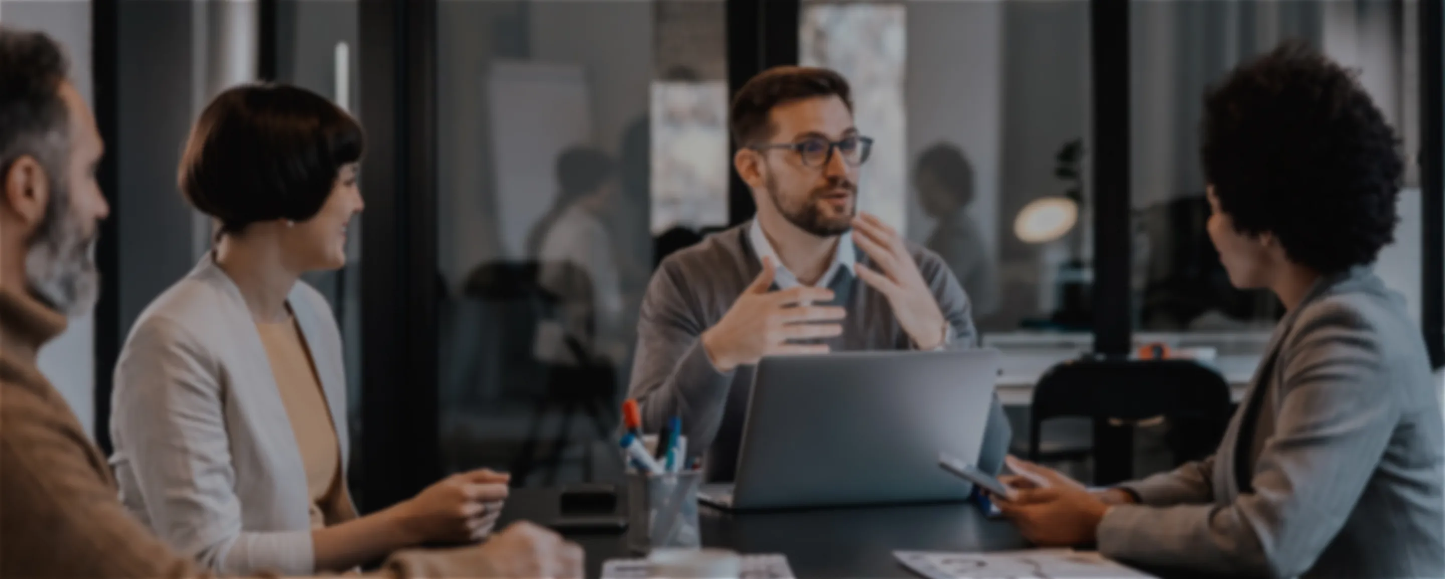 A group of coworkers sit around a conference table in an office, talking and looking at a laptop and tablet during a meeting.