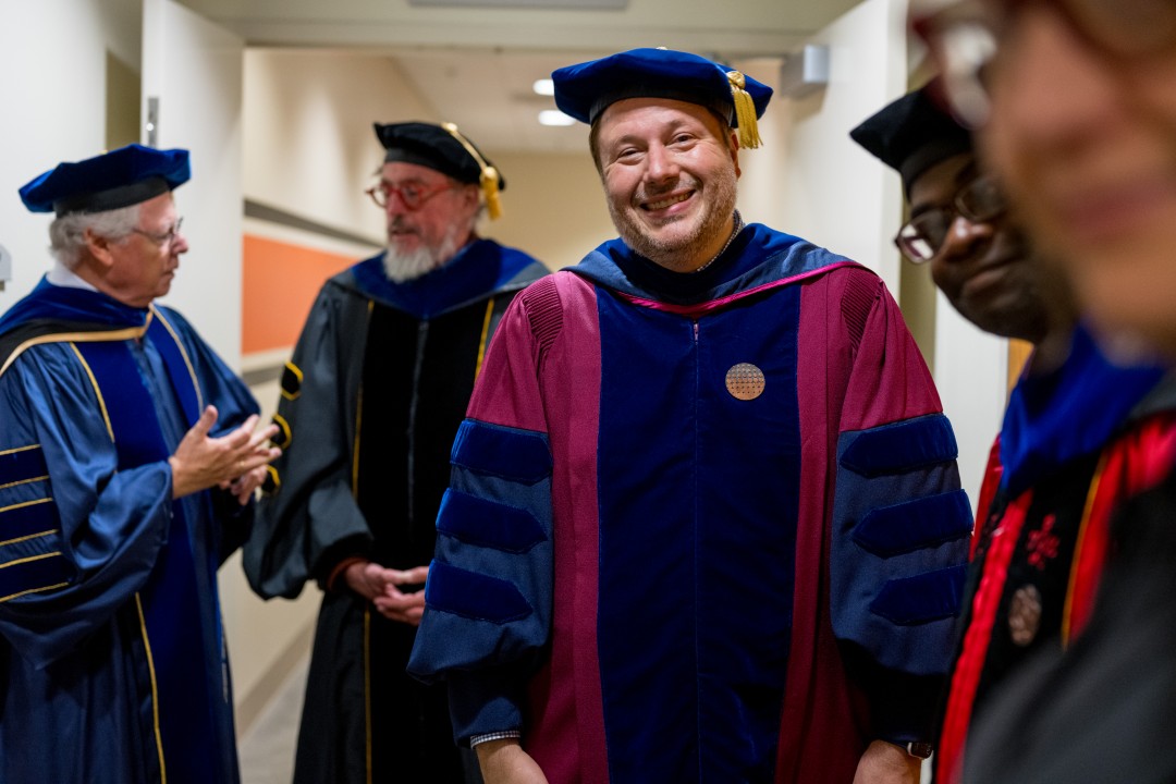 A smiling faculty member in colorful academic robes stands backstage with colleagues before the procession.