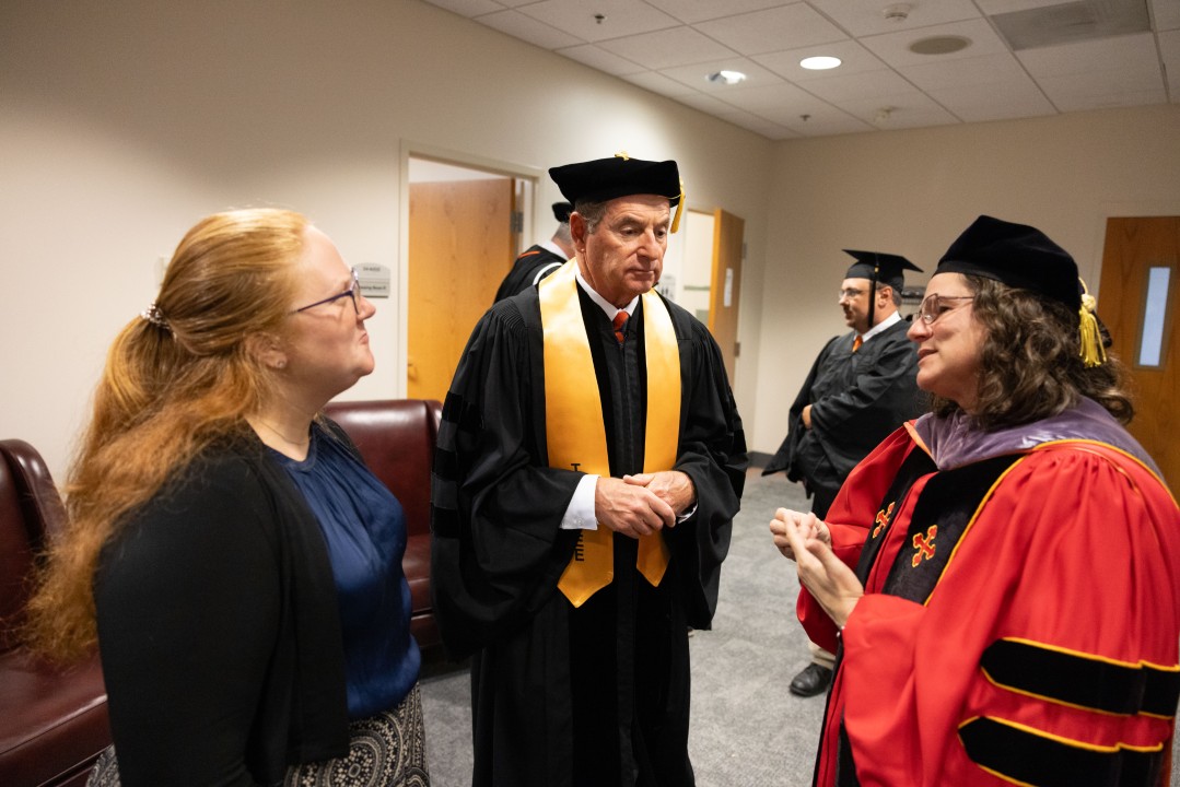 Faculty and staff chat together backstage, with one professor in regalia speaking with two colleagues.