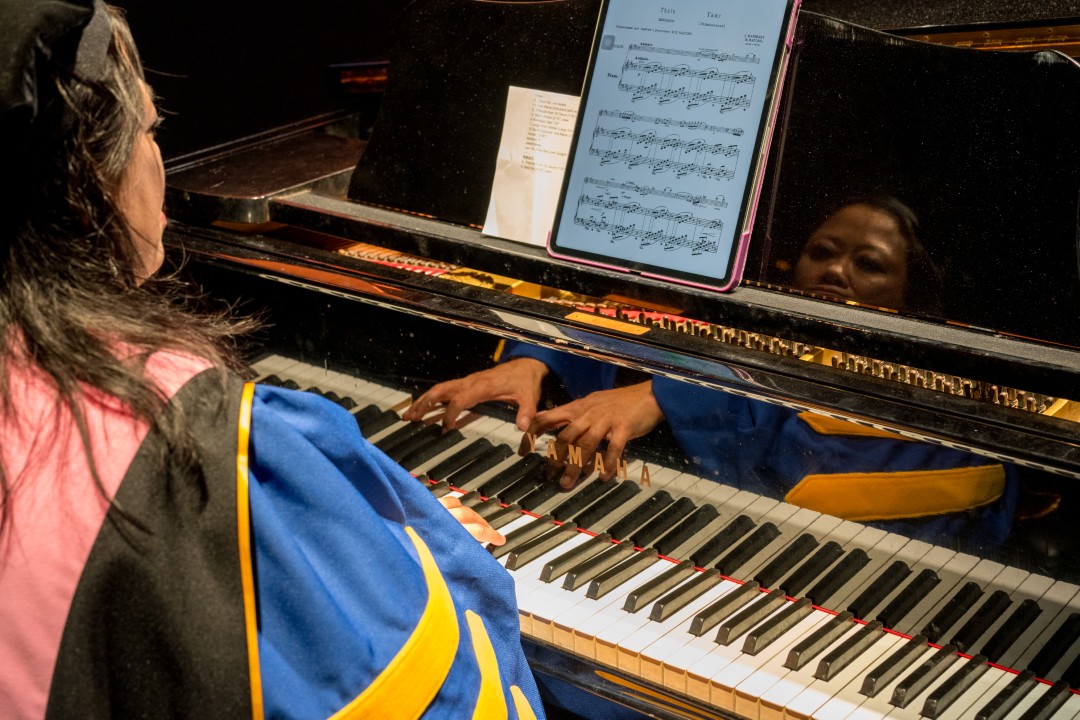 A faculty member in academic regalia plays piano with sheet music displayed on a tablet.