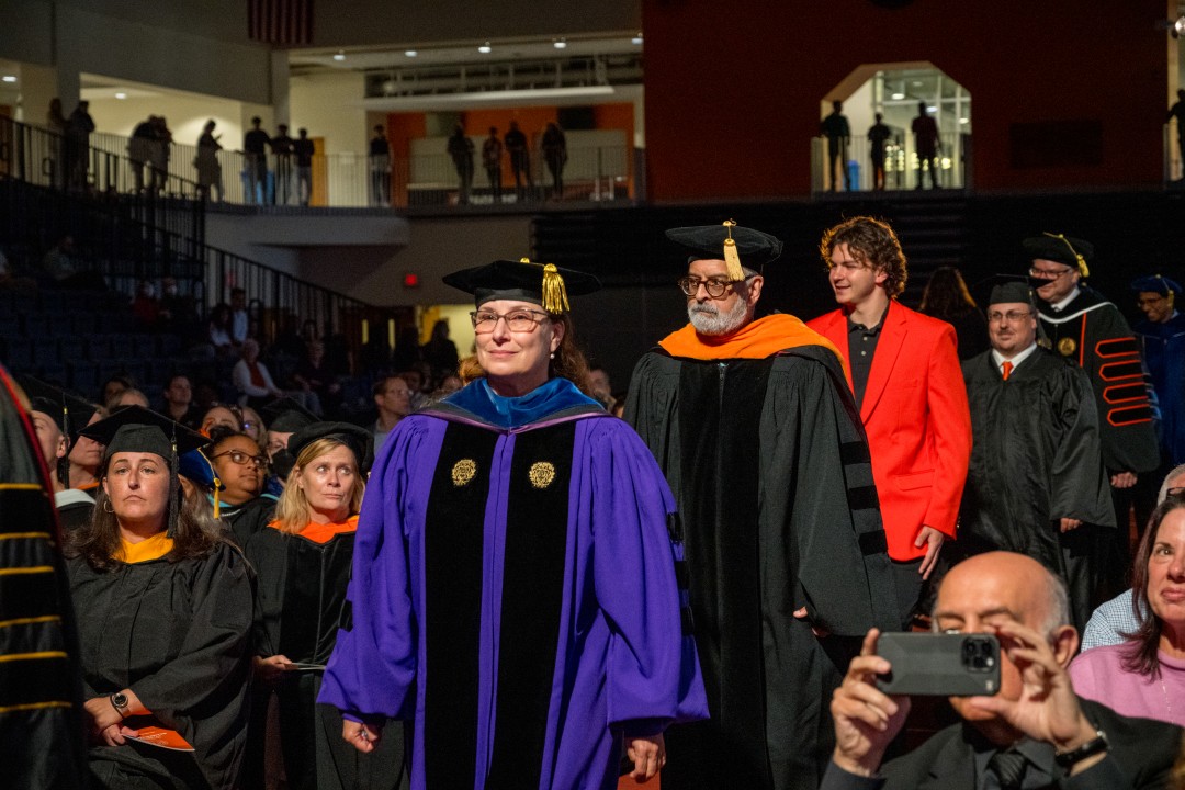 Faculty in colorful academic robes walk in the inauguration procession before a seated audience.