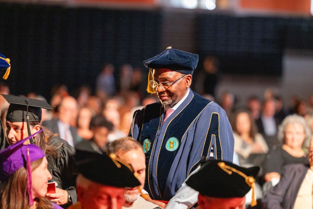A faculty member in a blue velvet gown and cap smiles while walking in the inauguration procession.