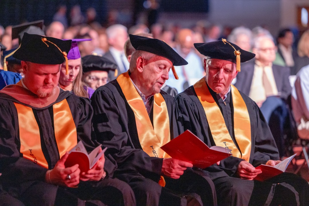 Faculty members wearing gold honor stoles read programs while seated at the ceremony.