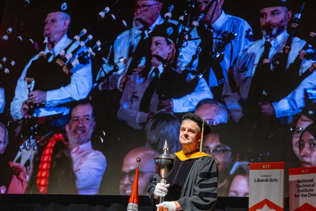 A faculty marshal in regalia holds a ceremonial mace in front of a video of bagpipers on a projector screen.