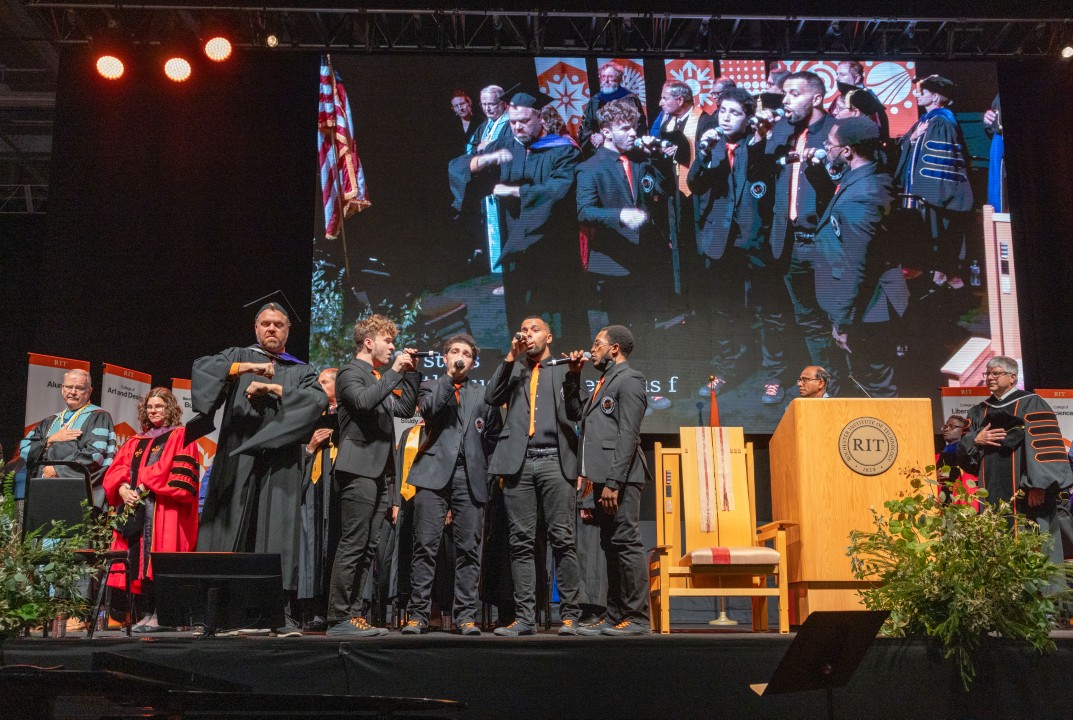 A student a cappella group performs on stage during the inauguration ceremony with a large screen behind them.