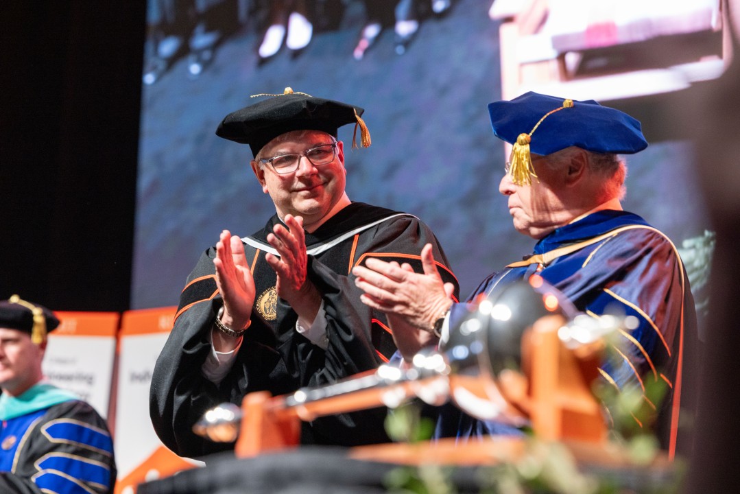 R I T president Sanders applauds on stage beside a colleague with the ceremonial mace in the foreground.