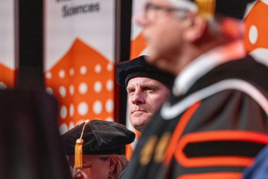 A faculty member in academic regalia looks forward with a serious expression as colleagues sit nearby.