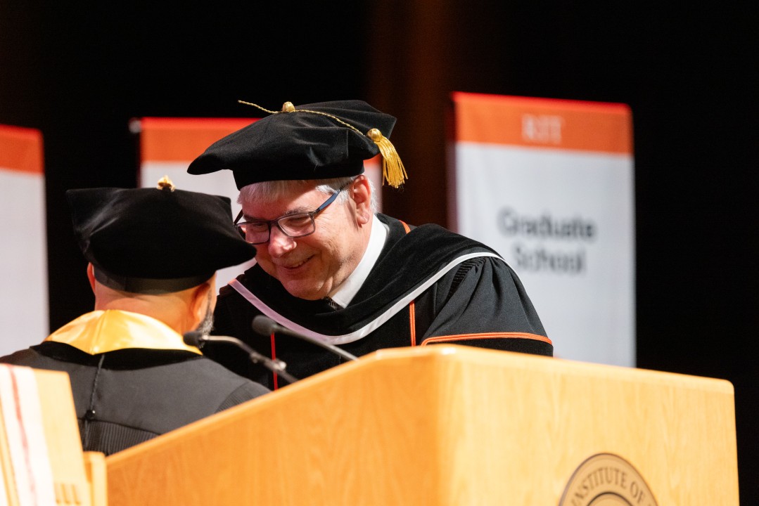 President Sanders in regalia smiles as he speaks with a colleague at the podium.