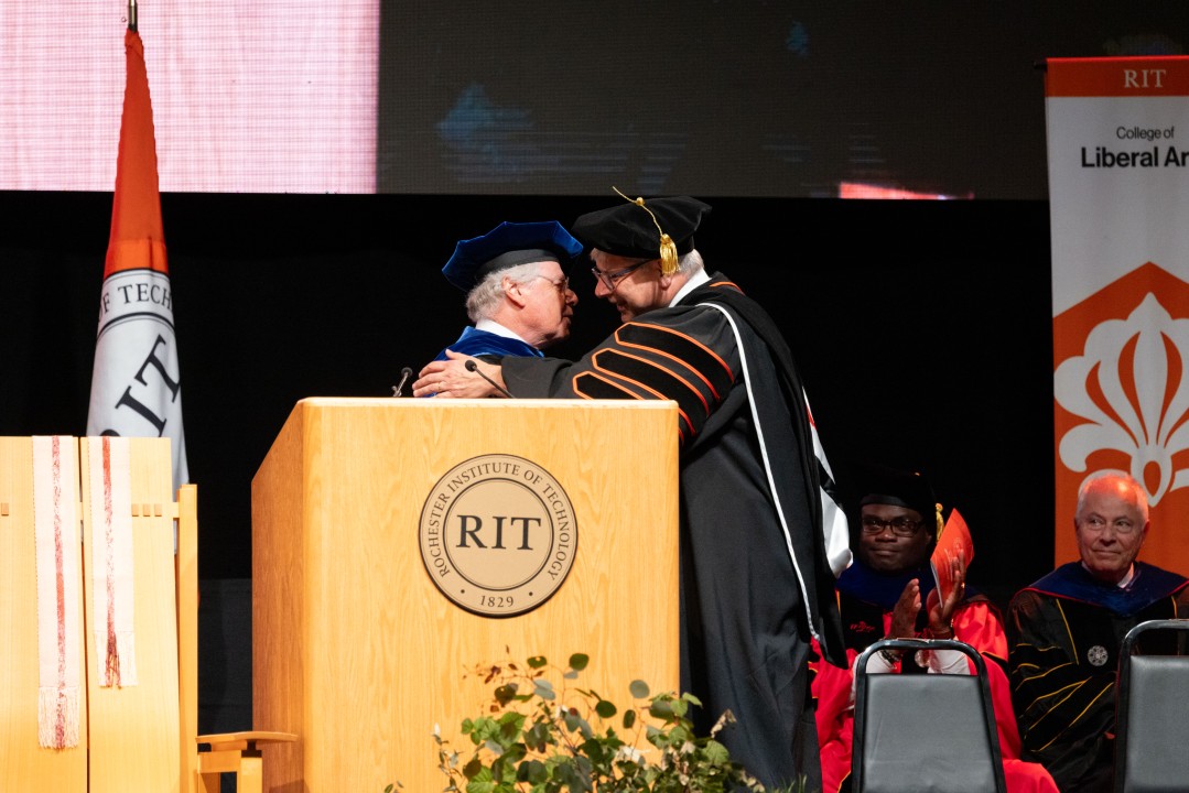 President Sanders hugs a guest speaker on stage beside the R I T podium.