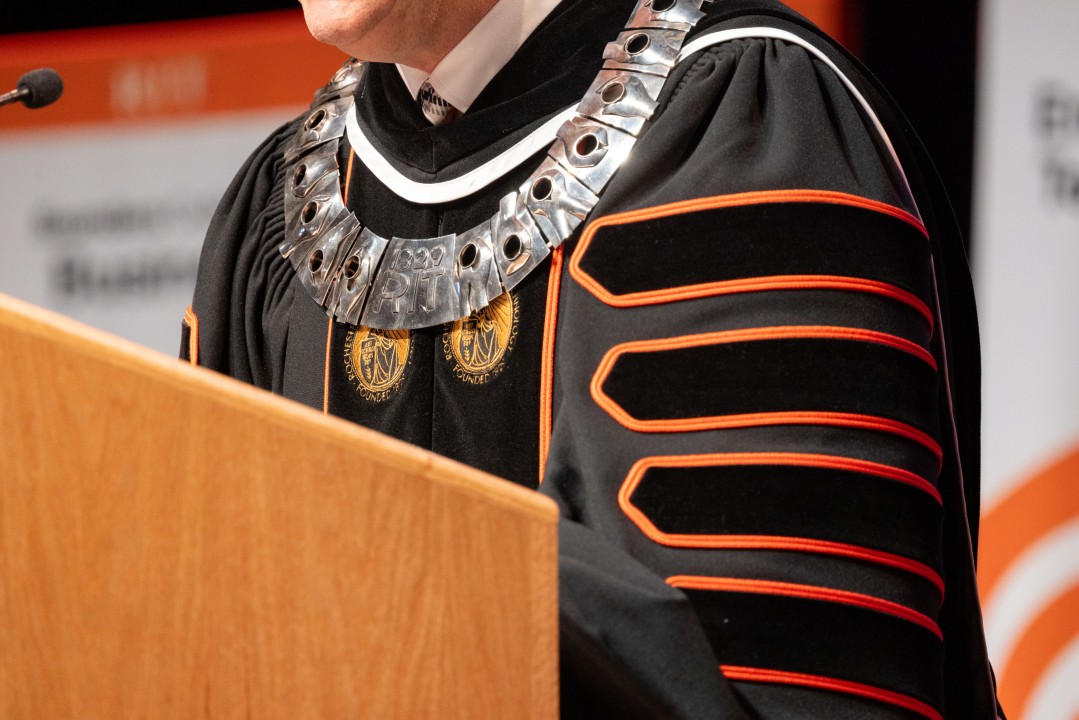 Close-up of president Sanders wearing the silver R I T presidential medallion over academic regalia while speaking at the podium.