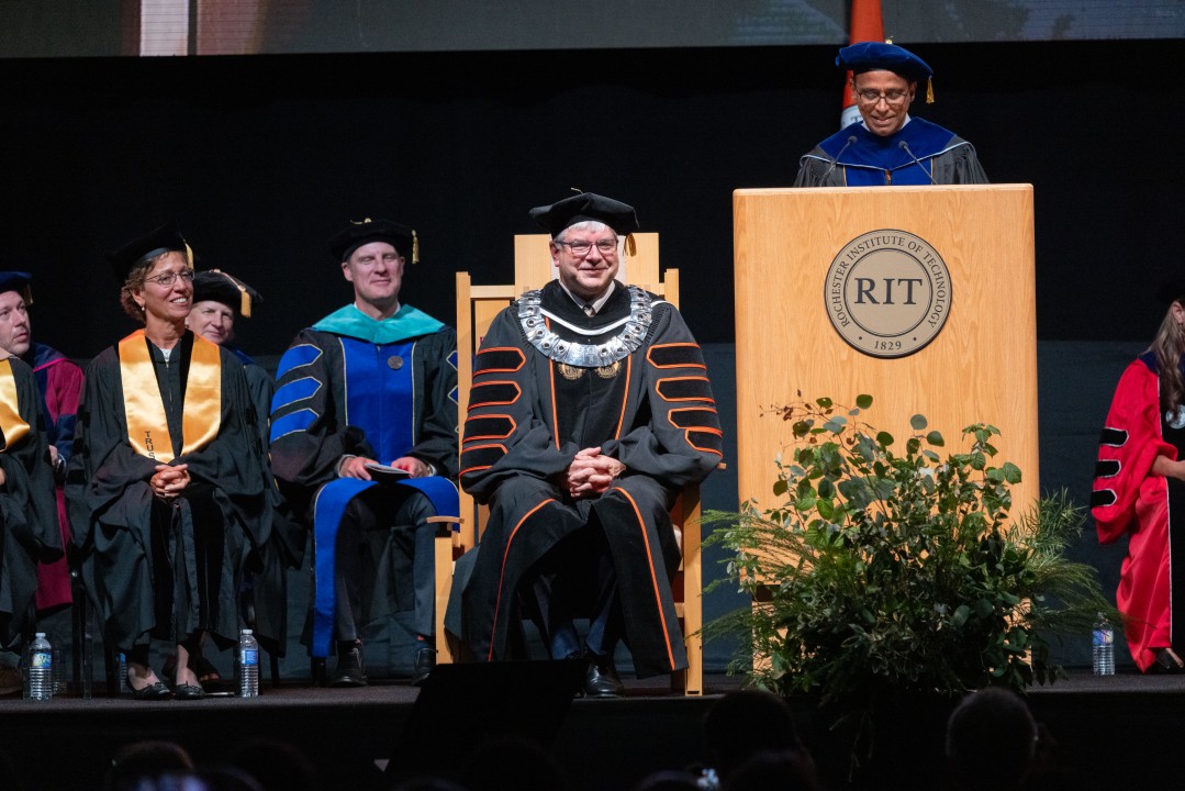 The newly inaugurated President Sanders sits smiling in regalia with a large medallion as a speaker addresses the audience at the podium.