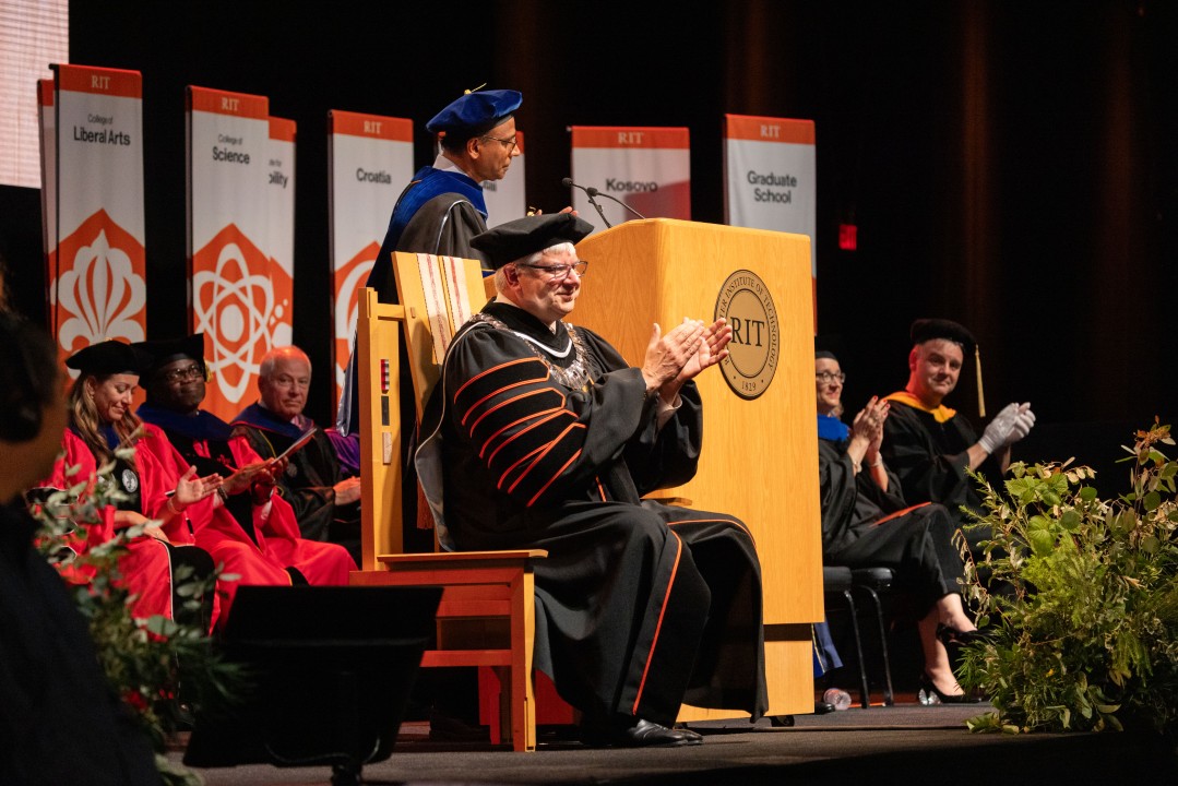 R I T President Sanders in regalia applauds on stage as a faculty speaker addresses the audience at the podium.